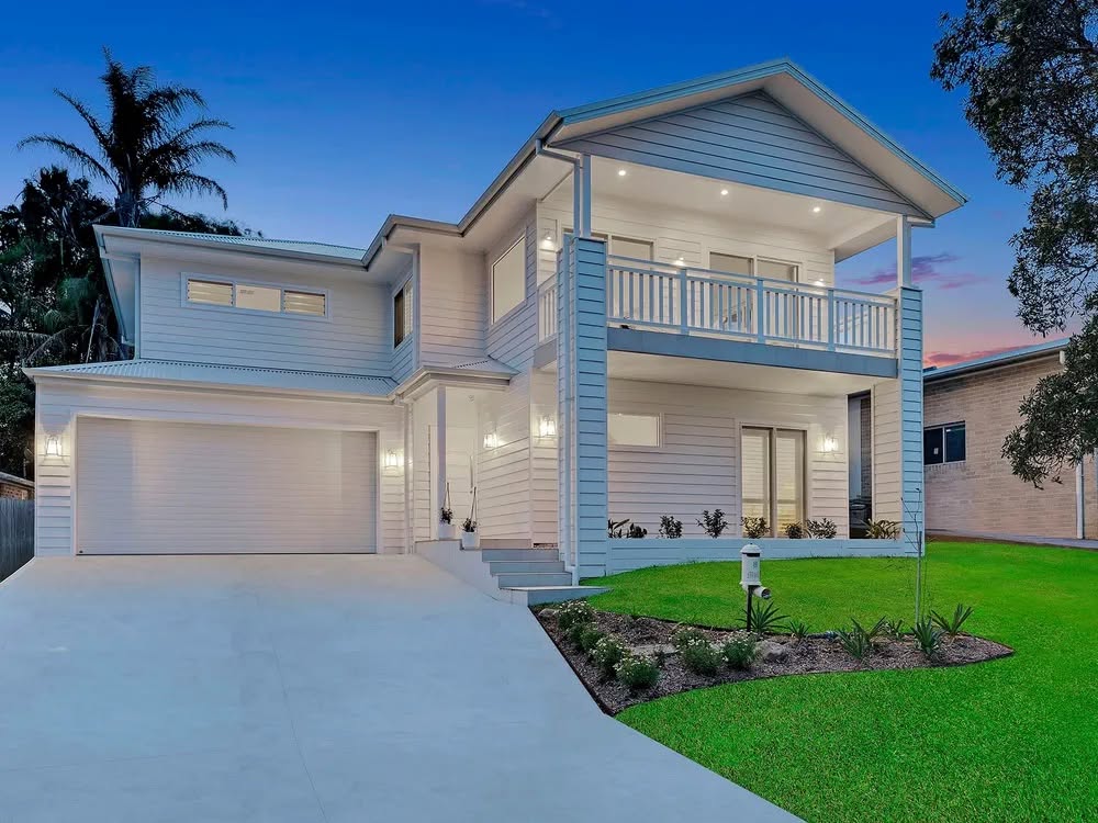 Two storey Hamptons style coastal home at dusk with balcony and double garage